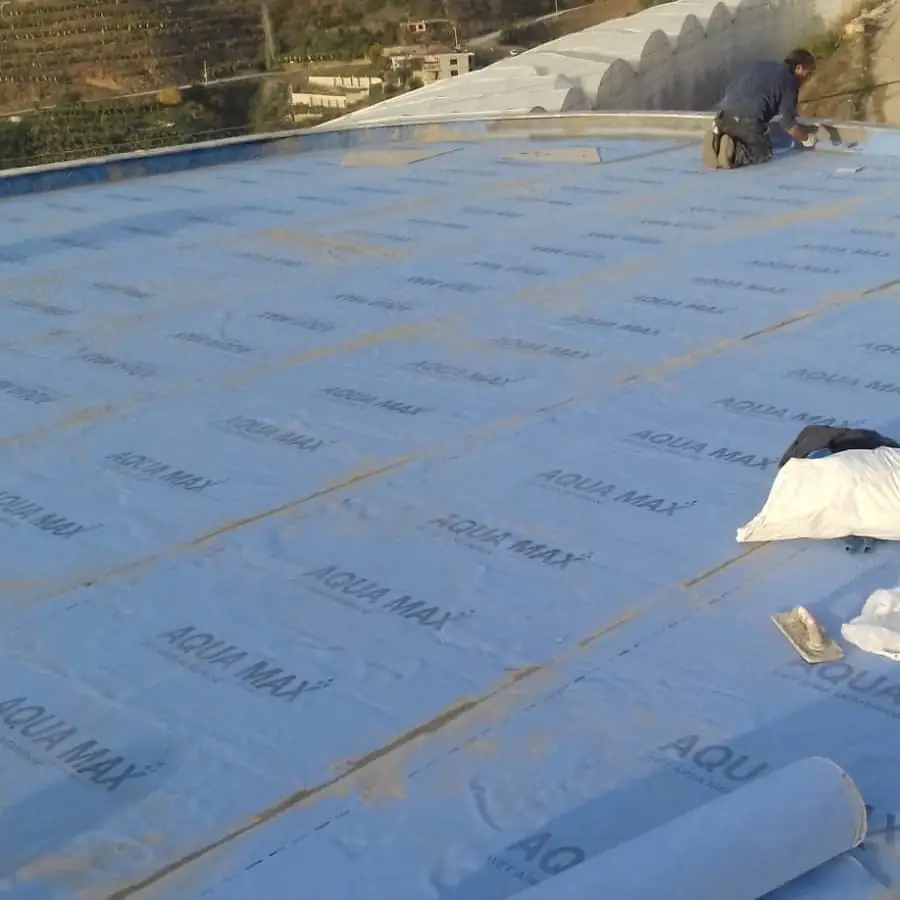 Teras çatıda serili mavi su yalıtım örtüsü ve parapet kenarında çalışan usta, önde mala