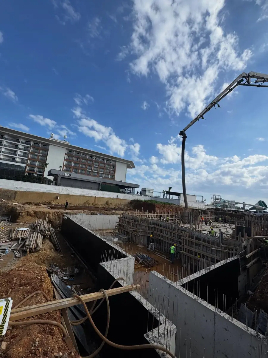 Temel kazısında beton pompasıyla döküm; solda su yalıtımı tamamlanmış perde duvarın siyah yüzeyi
